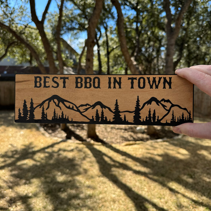 Hand holding a HipFlaskPlus Best BBQ in Town mountain scene black and natural wood sign outdoors under oak trees