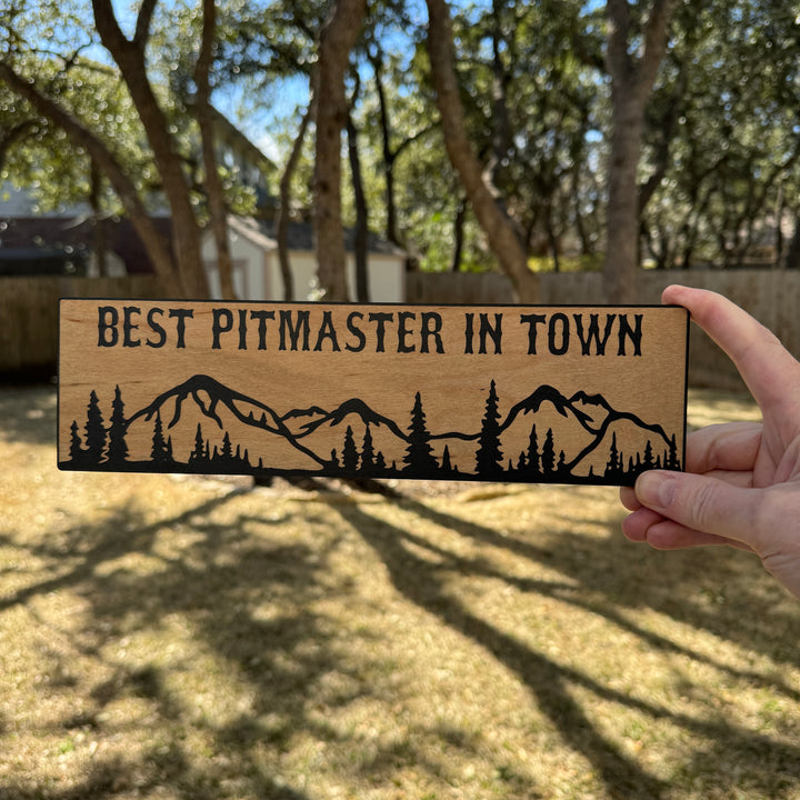 Hand holding a HipFlaskPlus Best Pitmaster in Town mountain scene black and natural wood sign outdoors under oak trees