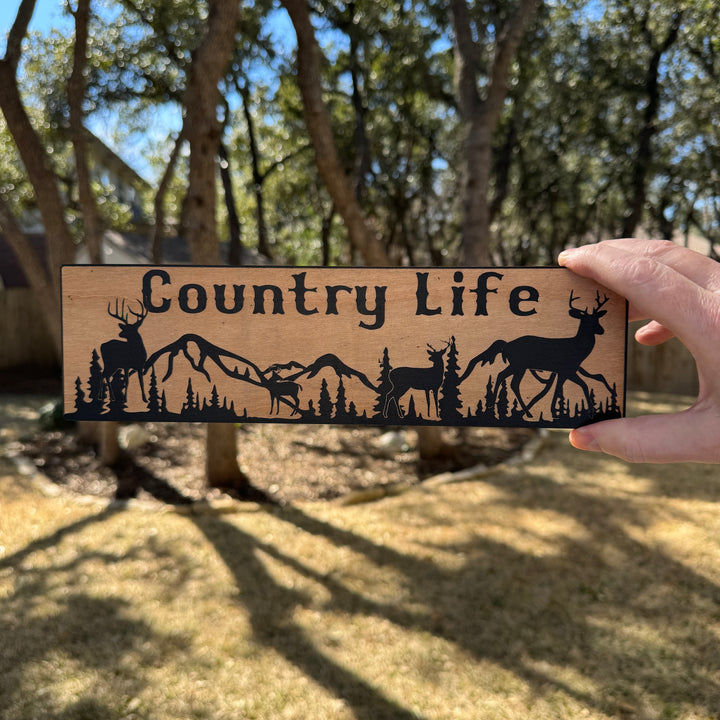 Hand holding a HipFlaskPlus Country Life deer mountain scene black and natural wood sign outdoors under oak trees