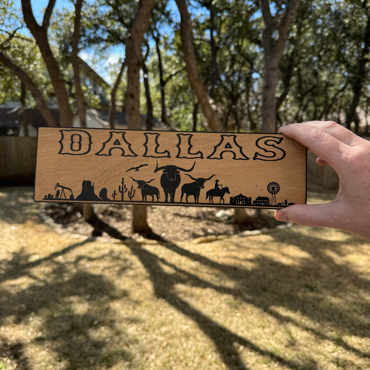 Hand holding a HipFlaskPlus Dallas landscape black and natural wood sign outdoors under oak trees