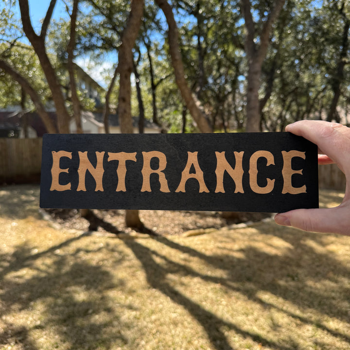 Hand holding a HipFlaskPlus Entrance black and natural wood sign outdoors under oak trees