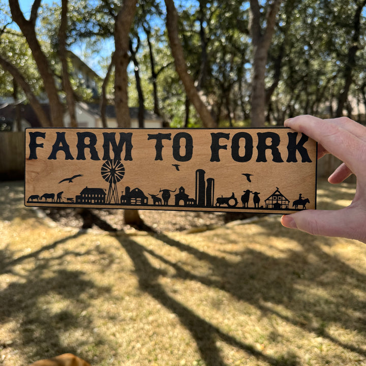Hand holding a HipFlaskPlus Farm to Fork black and natural wood sign outdoors under oak trees