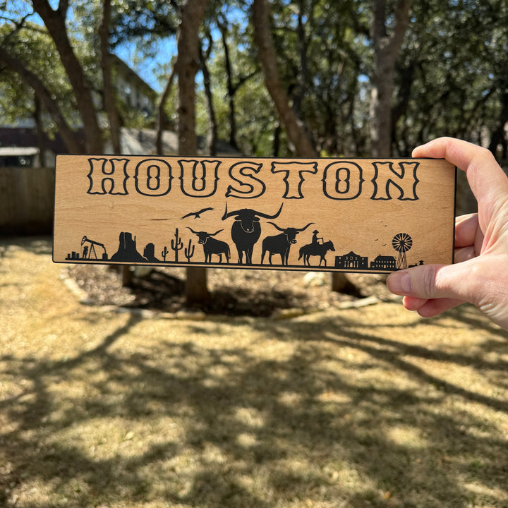 Hand holding a HipFlaskPlus Houston landscape black and natural wood sign outdoors under oak trees