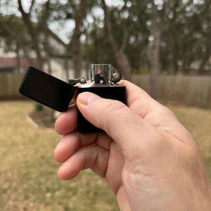 Hand holding an open HipFlaskPlus black windproof lighter in a backyard
