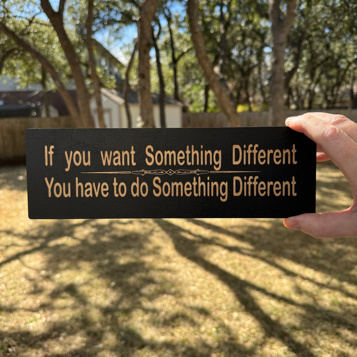 Hand holding a HipFlaskPlus If You Want Something Different You Have to Do Something Different black wood sign outdoors under oak trees