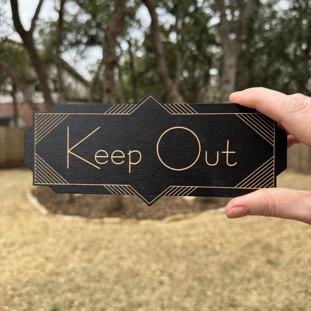 Hand holding a HipFlaskPlus Keep Out black art deco wood sign outdoors under oak trees