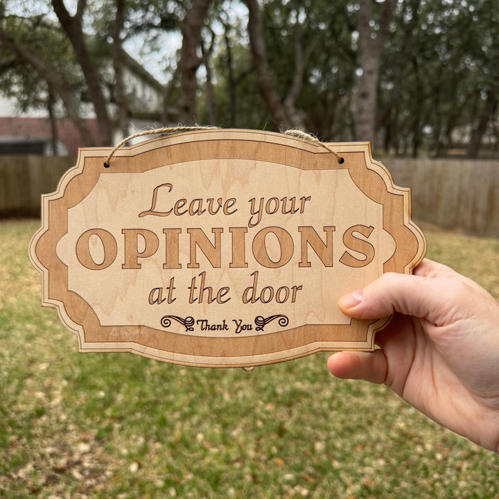 Hand holding a HipFlaskPlus Leave Your Opinions at the Door raw wood door sign in a garden