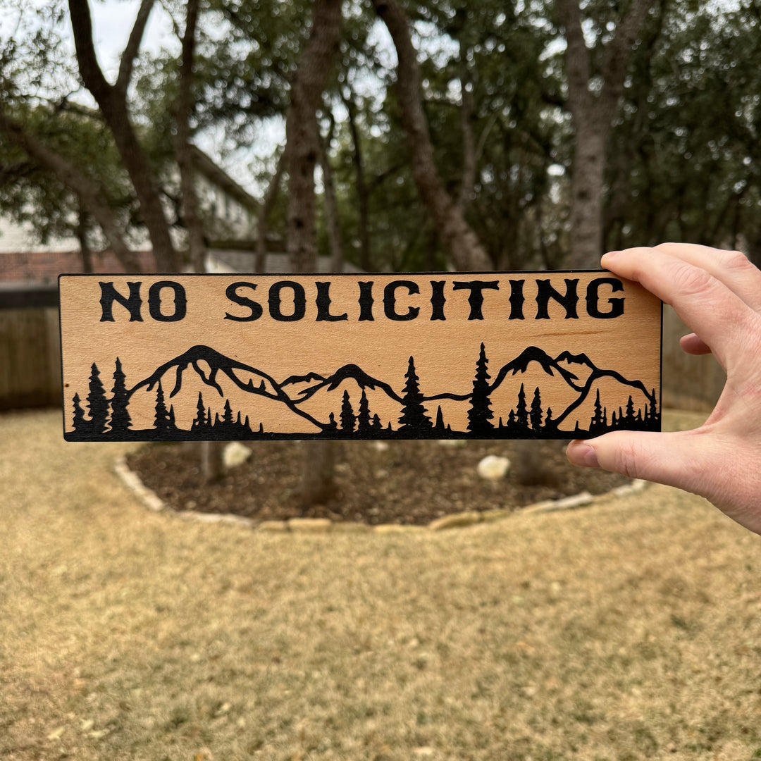Hand holding a HipFlaskPlus No Soliciting mountain scene black and natural wood sign outdoors under oak trees