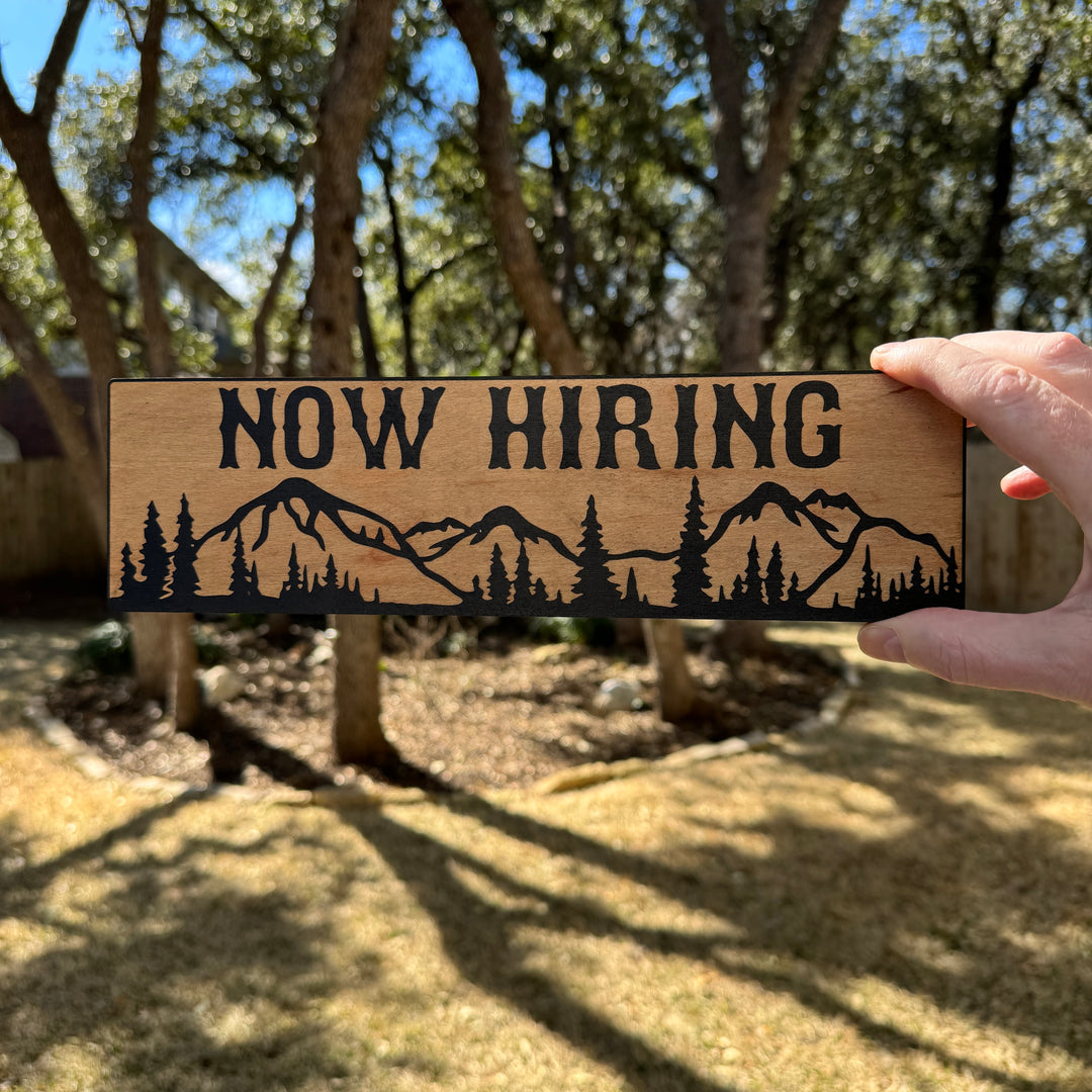 Hand holding a HipFlaskPlus Now Hiring mountain scene black and natural wood sign outdoors under oak trees