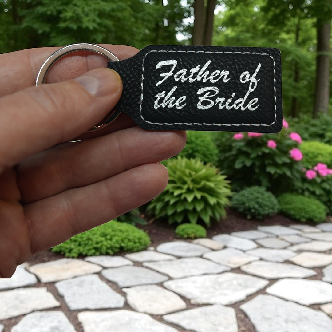 Hand holding a HipFlaskPlus black leather rectangle keychain with Father of the Bride engraving by a garden path