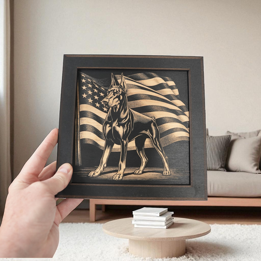 Hand holding a HipFlaskPlus Doberman and American flag engraved wood wall art in a black frame in a living room