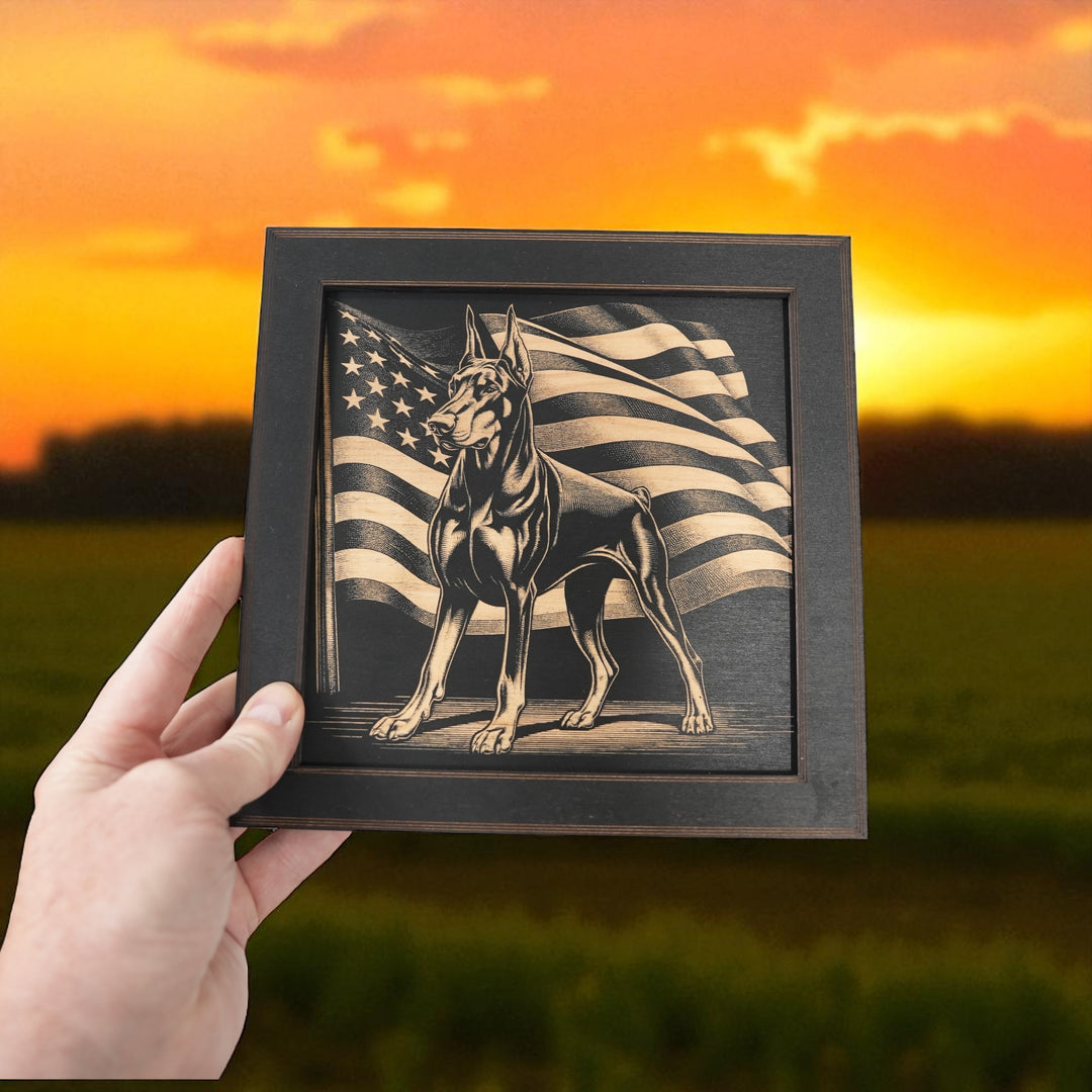 Hand holding a HipFlaskPlus Doberman and American flag engraved wood wall art in a black frame in a field at sunset
