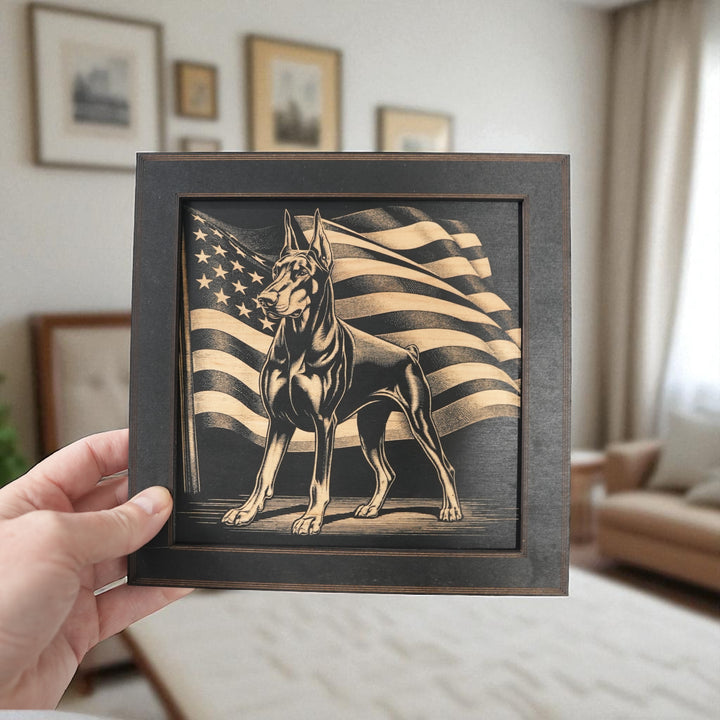Hand holding a HipFlaskPlus Doberman and American flag engraved wood wall art in a black frame in a living room