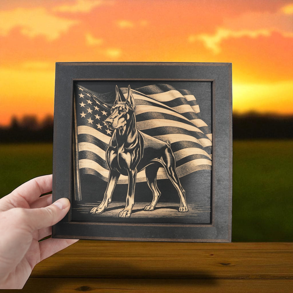 Hand holding a HipFlaskPlus Doberman and American flag engraved wood wall art in a black frame in a field at sunset