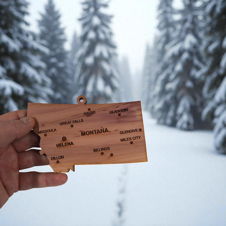 Hand holding a HipFlaskPlus custom Montana state ornament with cities in a snowy landscape
