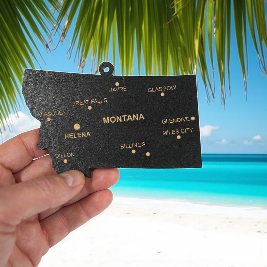 Hand holding a HipFlaskPlus custom Montana state ornament with cities on a tropical beach
