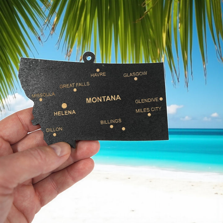 Hand holding a HipFlaskPlus custom Montana state ornament with cities on a tropical beach