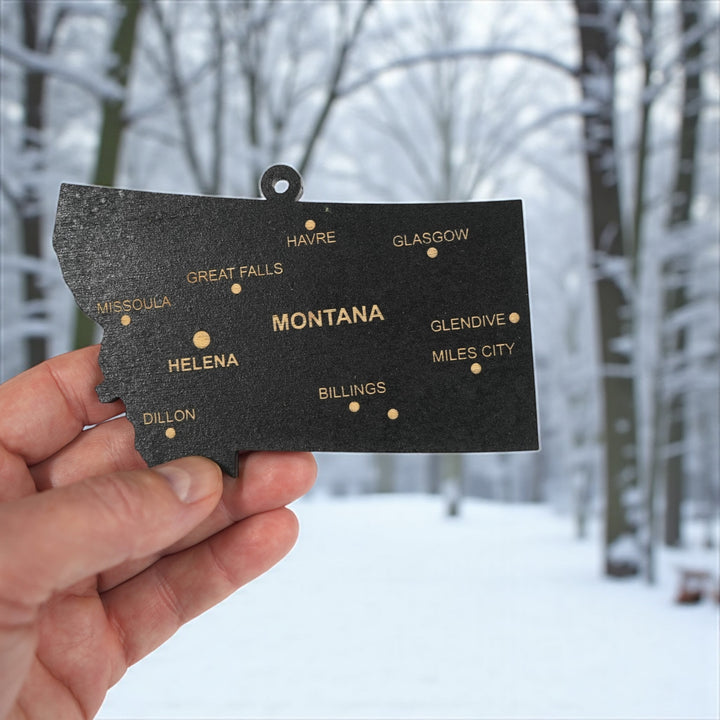 Hand holding a HipFlaskPlus custom Montana state ornament with cities in a snowy landscape