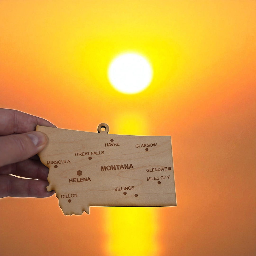 Hand holding a HipFlaskPlus custom Montana state ornament with cities at sunset