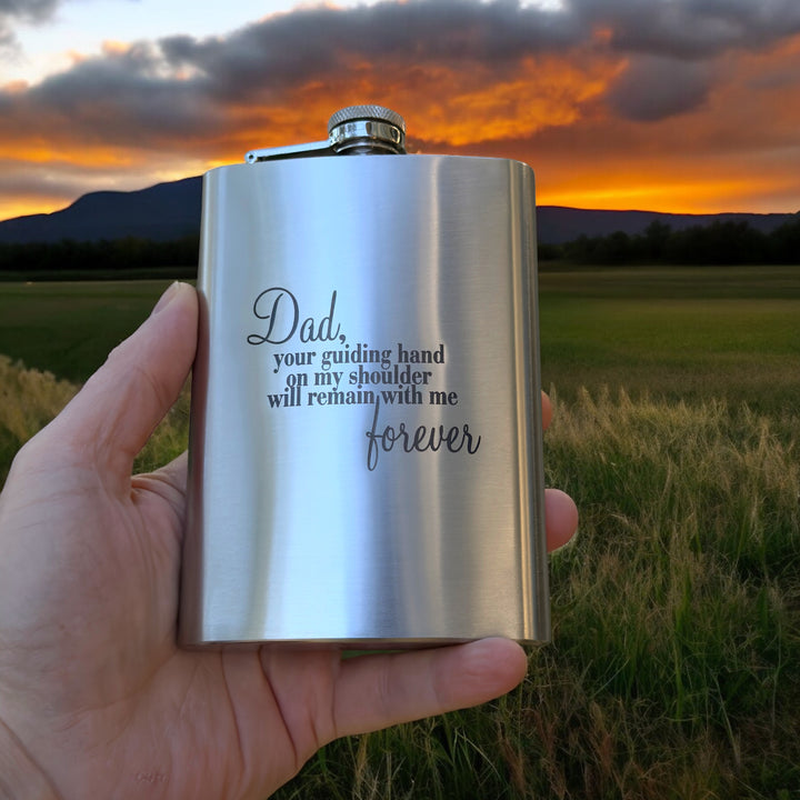 Hand holding a HipFlaskPlus 8oz stainless steel hip flask engraved with Dad Your Guiding Hand poem in a field at sunset