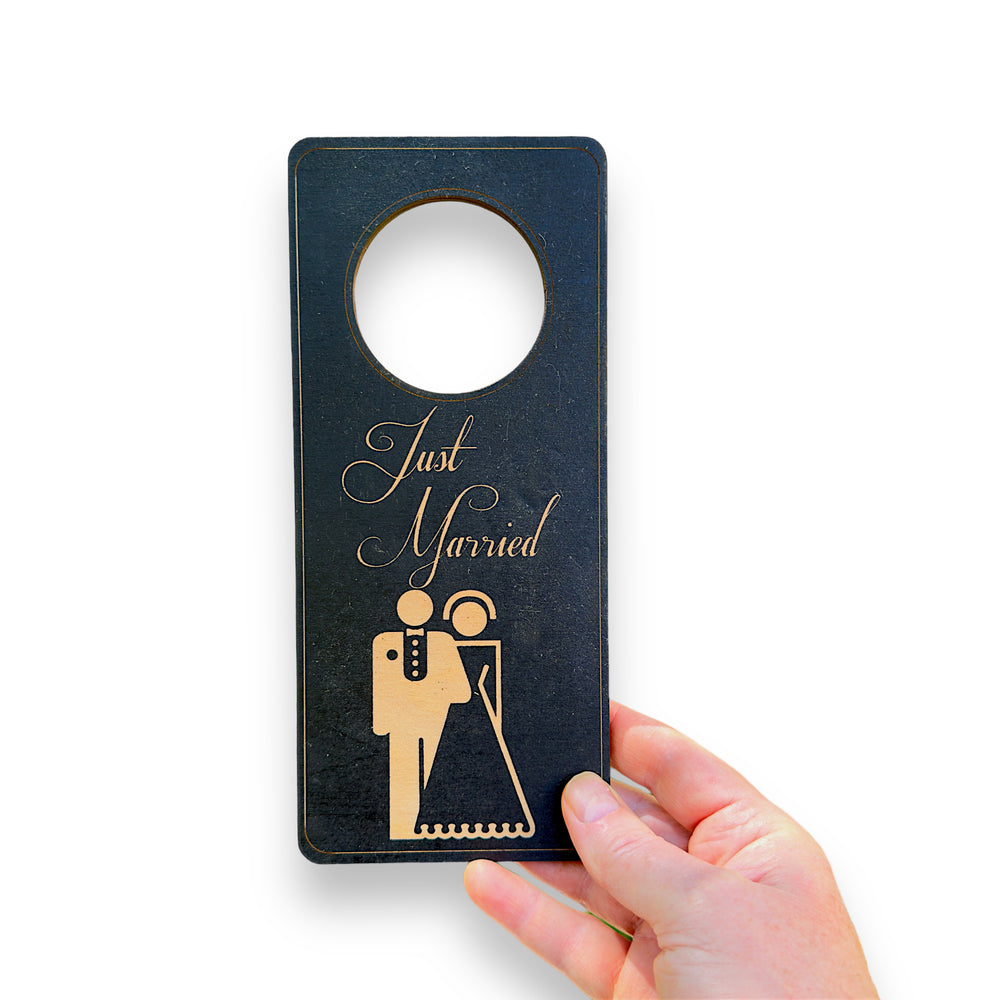 Hand holding a HipFlaskPlus black wood door hanger engraved with Just Married with a bride and groom