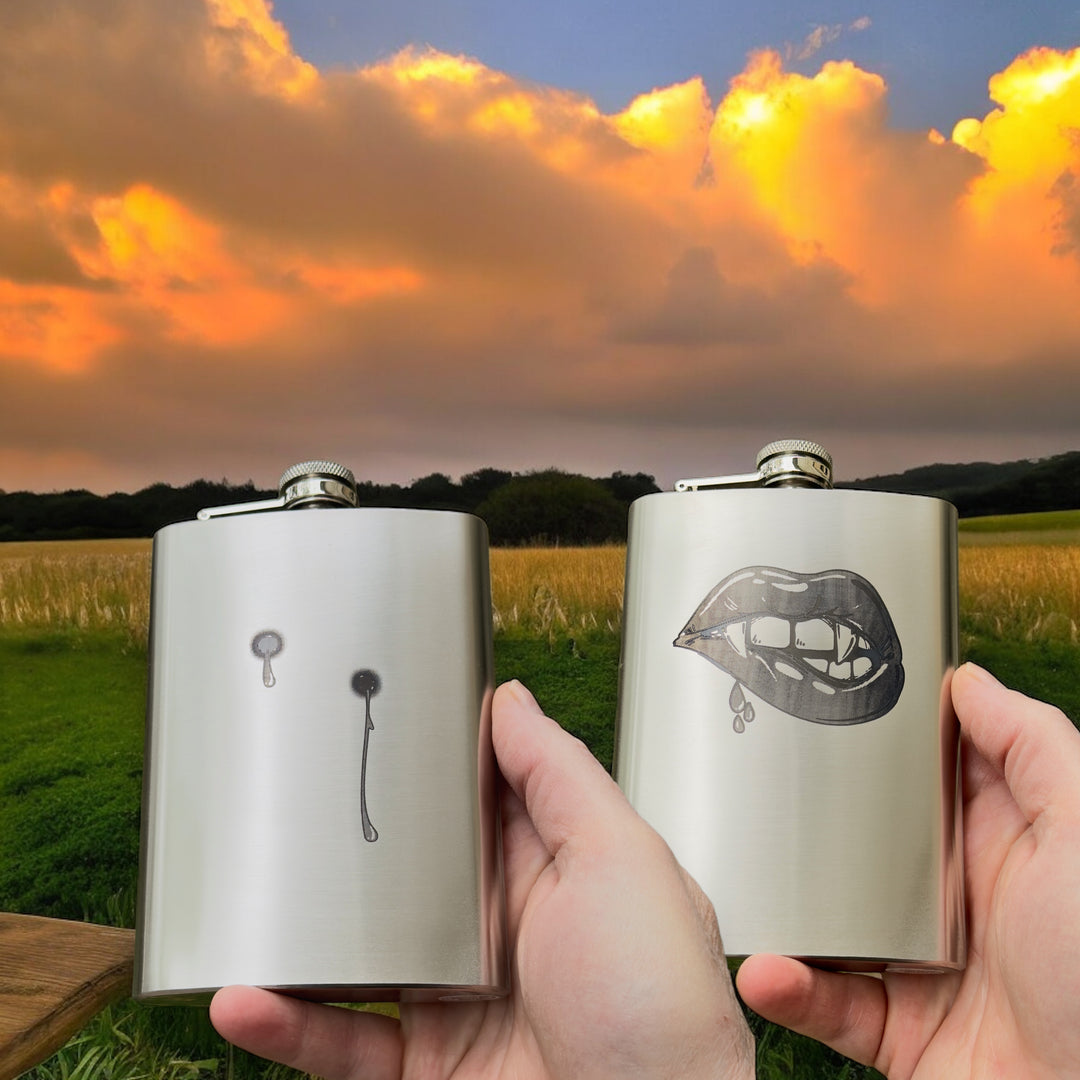 Hand holding HipFlaskPlus 8oz stainless steel hip flasks engraved with his and hers vampire lips and fangs at sunset