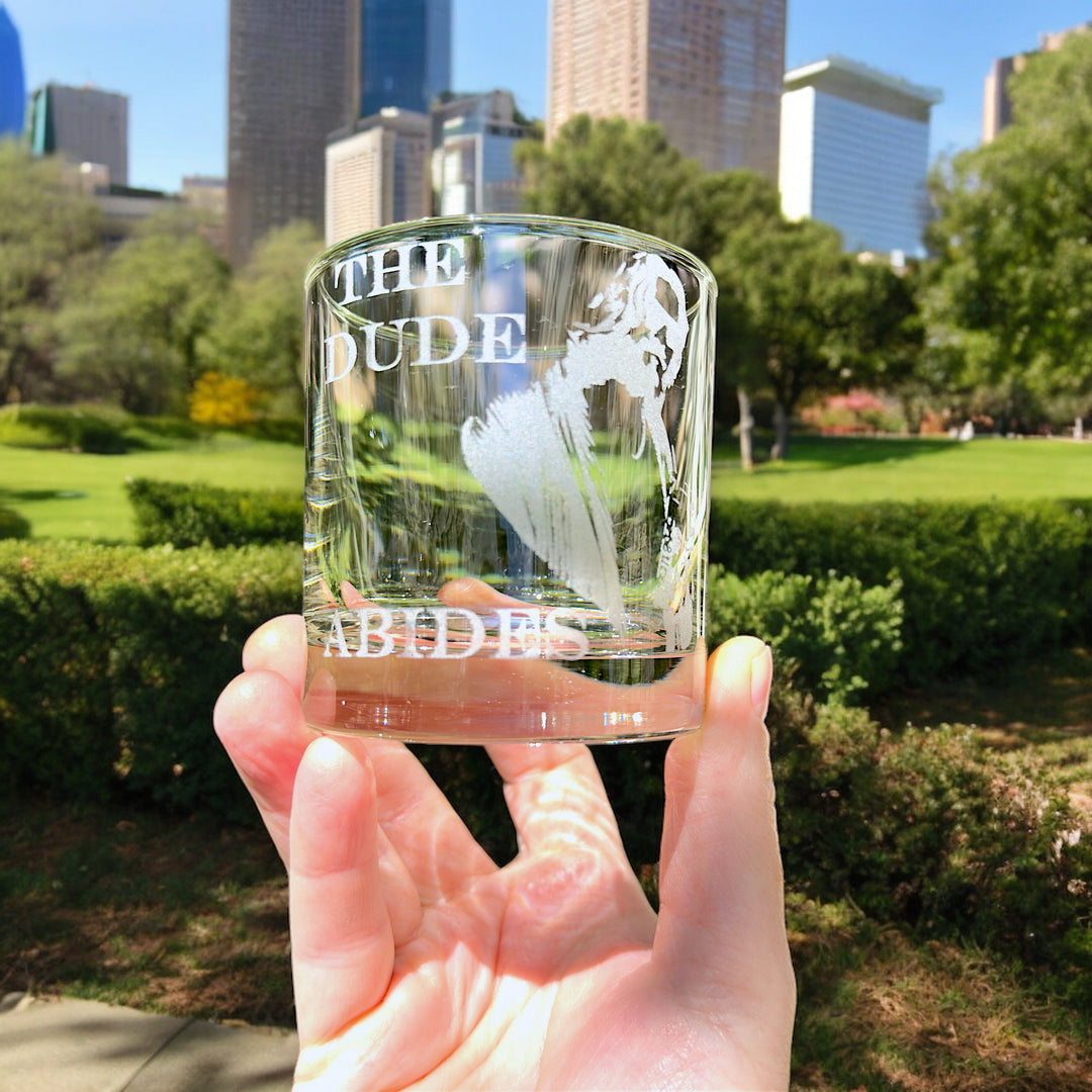Hand holding a HipFlaskPlus rocks glass engraved with The Dude Abides in a city park