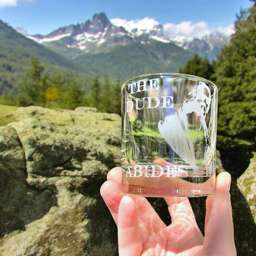 Hand holding a HipFlaskPlus rocks glass engraved with The Dude Abides with snowy mountains in the background