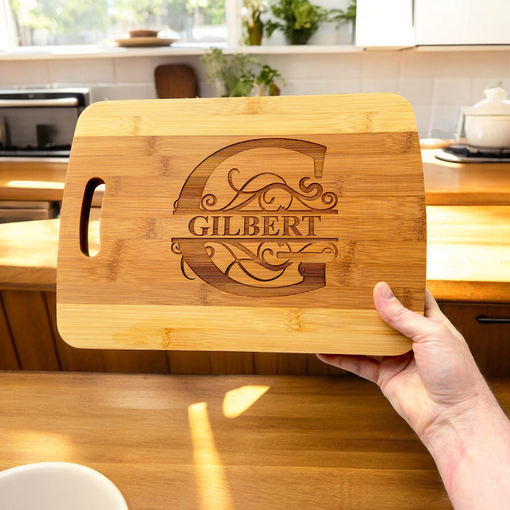 Hand holding a HipFlaskPlus bamboo cutting board engraved with a personalized split letter monogram and name in a kitchen