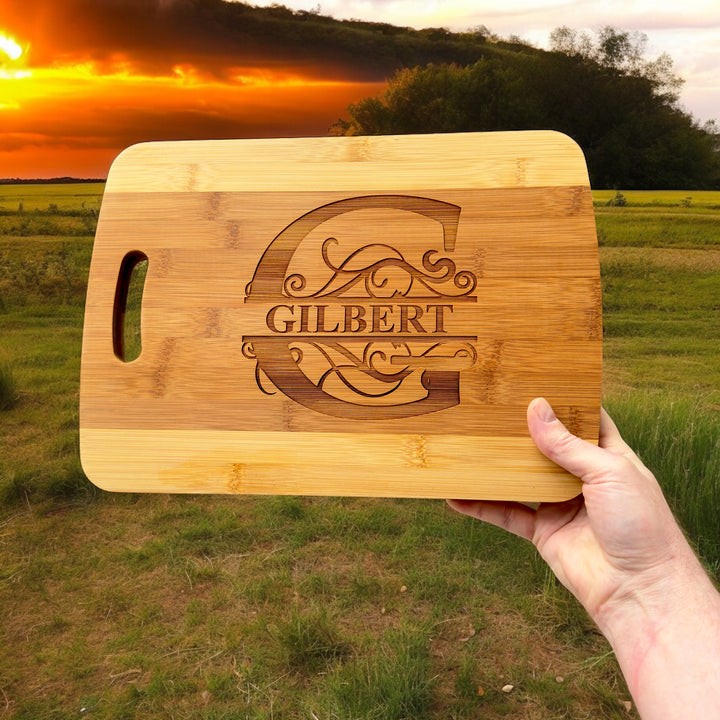 Hand holding a HipFlaskPlus bamboo cutting board engraved with a personalized split letter monogram and name at sunset over a field