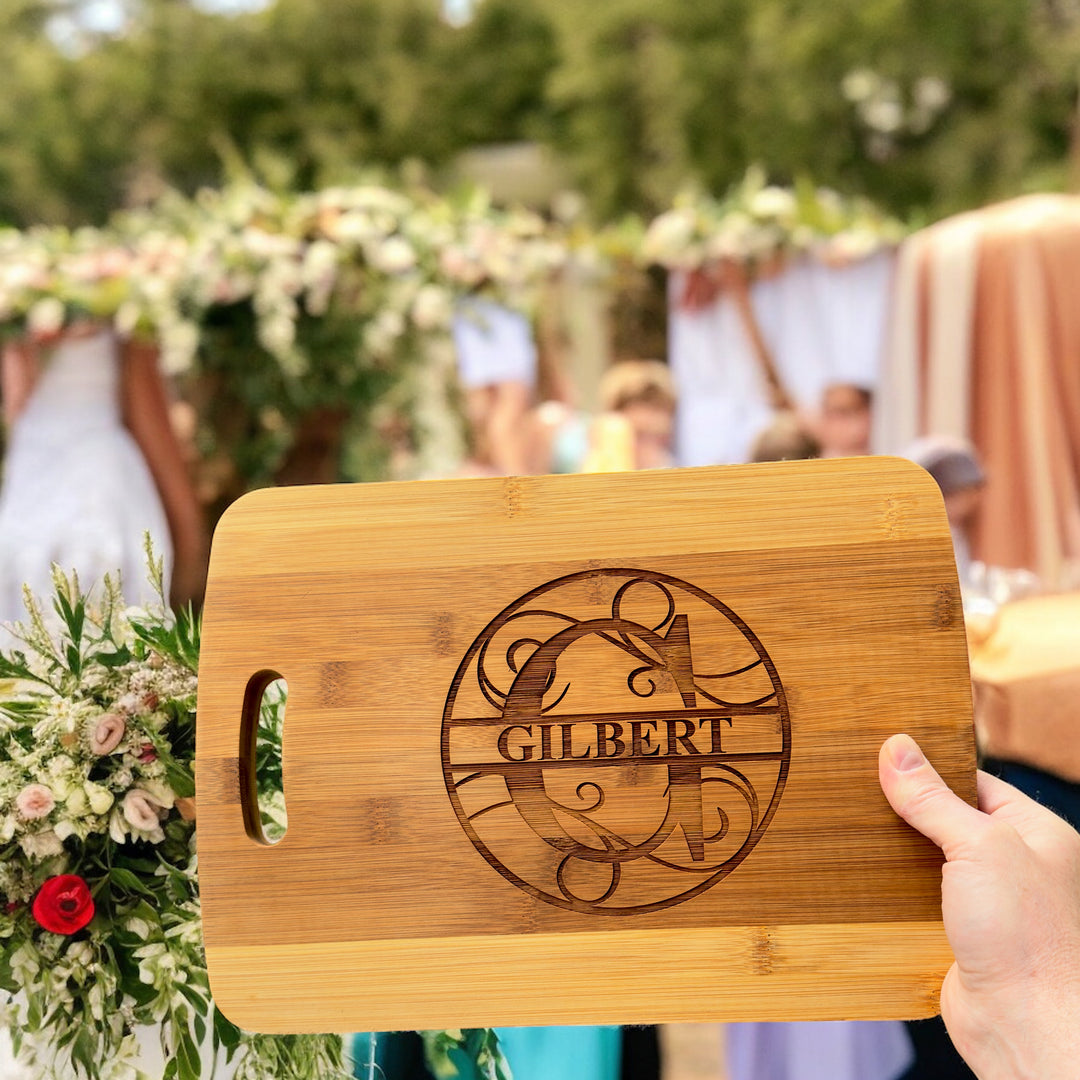 Hand holding a HipFlaskPlus bamboo cutting board engraved with a personalized split letter monogram and name at a wedding with flowers