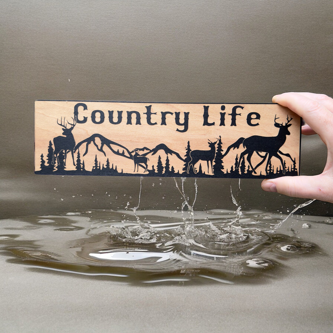 Hand holding a HipFlaskPlus Country Life deer mountain scene black and natural wood sign by driftwood