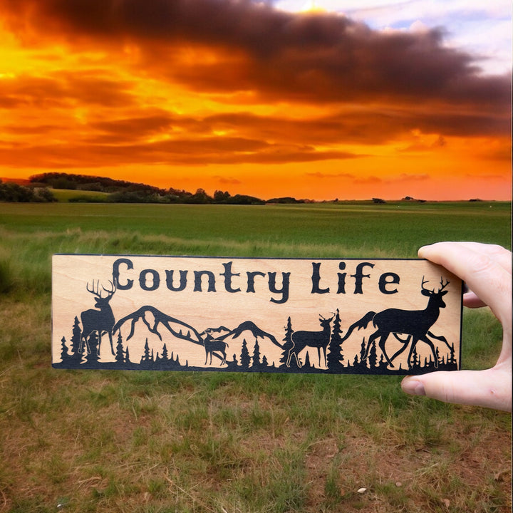 Hand holding a HipFlaskPlus Country Life deer mountain scene black and natural wood sign at sunset