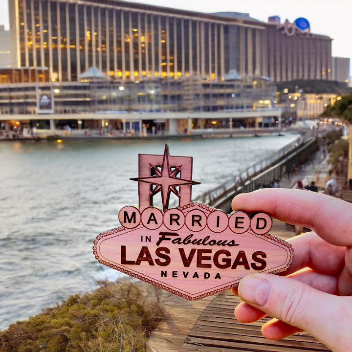 Hand holding a HipFlaskPlus cedar wood Married in Fabulous Las Vegas sign ornament by a waterfront hotel
