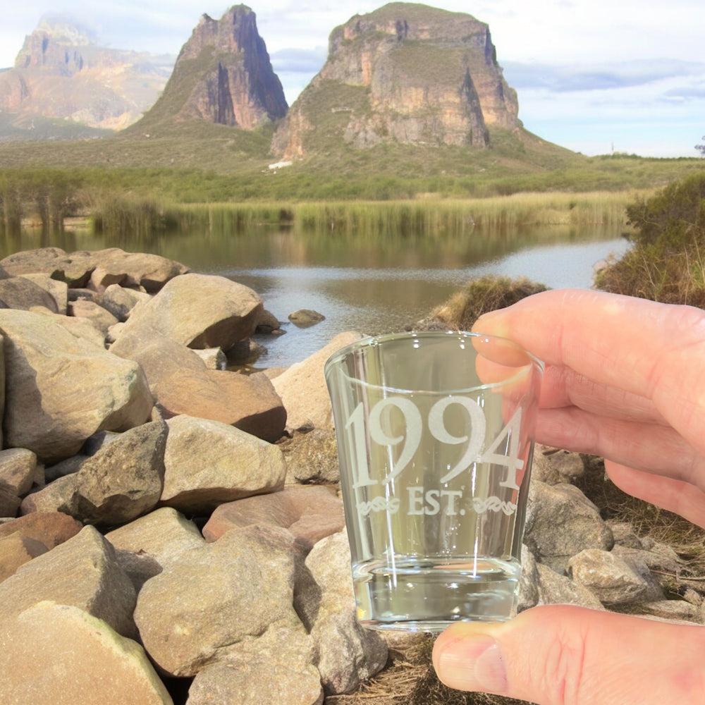 Hand holding a HipFlaskPlus Est. 1994 engraved shot glass by a lake with rock formations