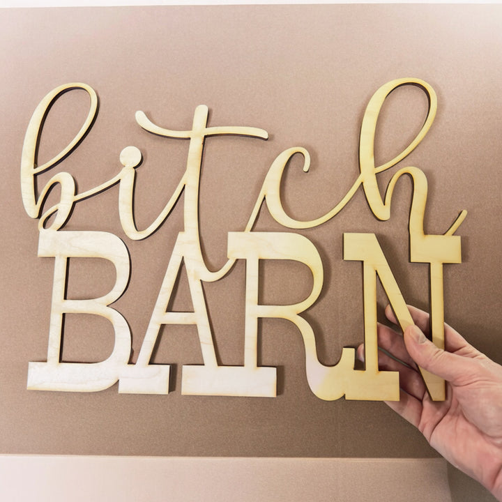 Hand holding a HipFlaskPlus Bitch Barn maple wood cutout sign on a gray background