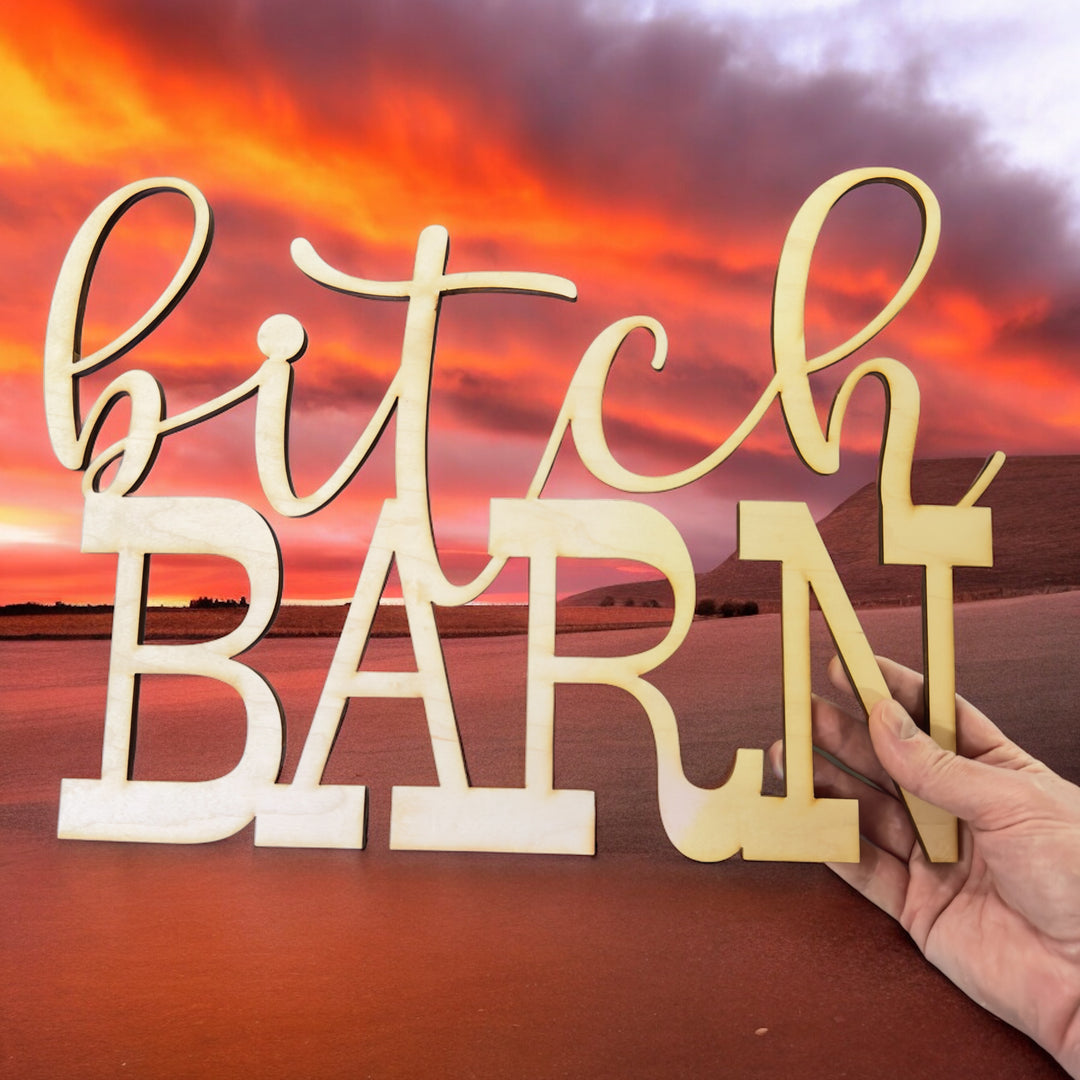 Hand holding a HipFlaskPlus Bitch Barn maple wood cutout sign at sunset