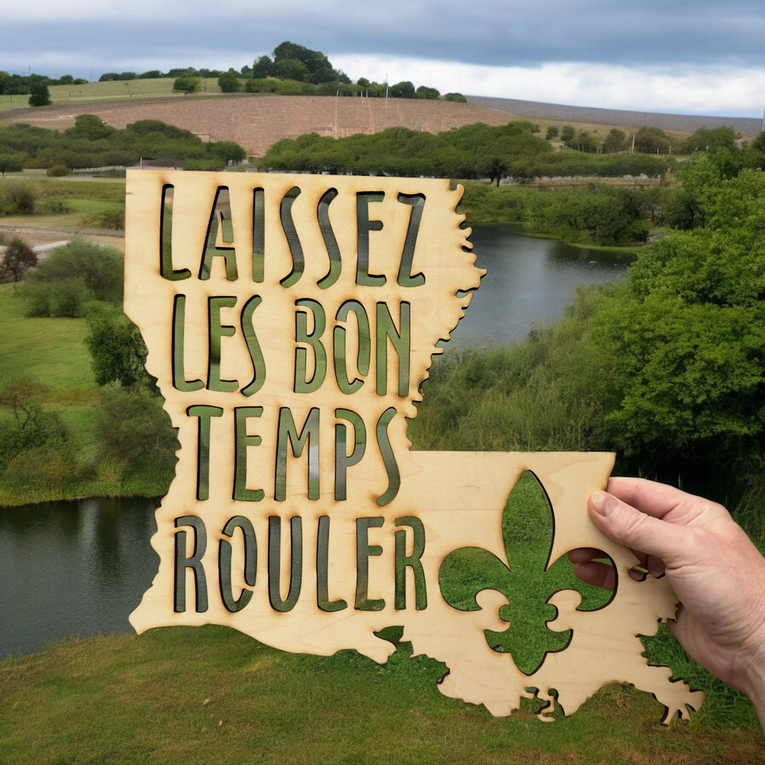 Hand holding a HipFlaskPlus Laissez les Bon Temps Rouler Louisiana maple wood cutout sign by a meadow