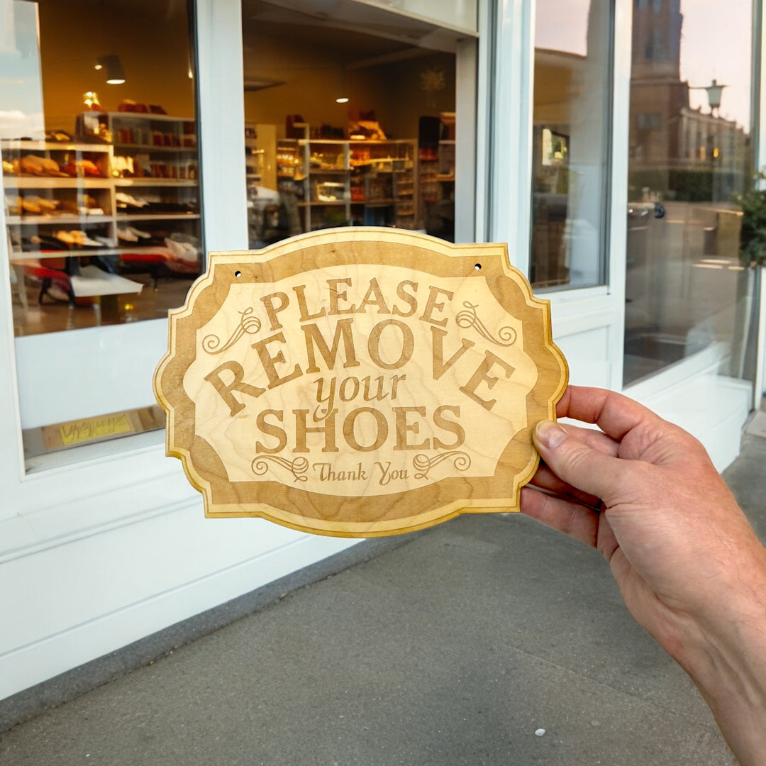 Hand holding a HipFlaskPlus Please Remove Your Shoes raw wood door sign in a shop doorway