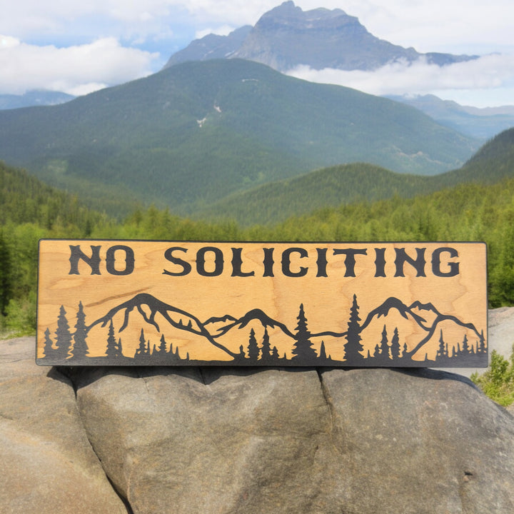 Hand holding a HipFlaskPlus No Soliciting mountain scene black and natural wood sign by mountains
