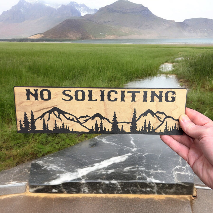 Hand holding a HipFlaskPlus No Soliciting mountain scene black and natural wood sign by mountains