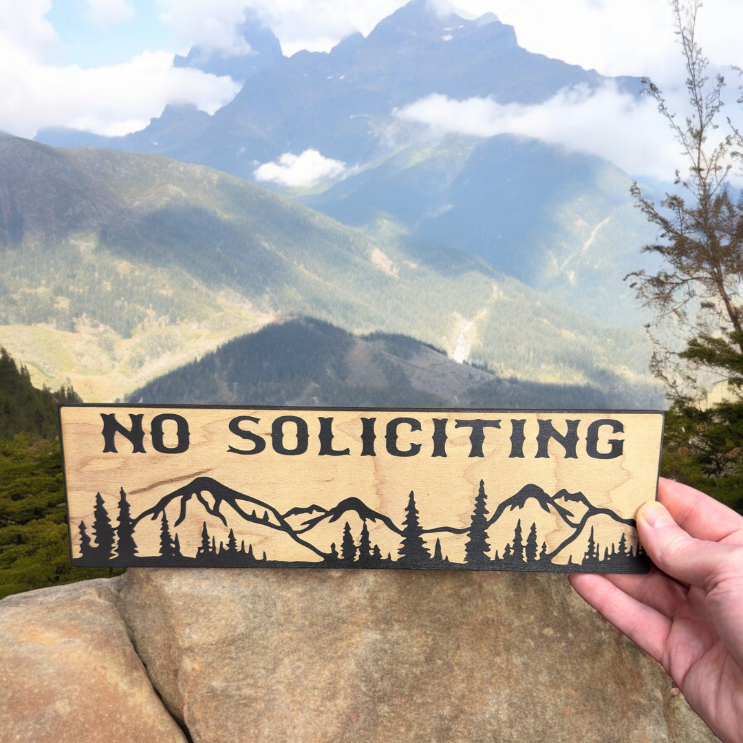 Hand holding a HipFlaskPlus No Soliciting mountain scene black and natural wood sign by mountains