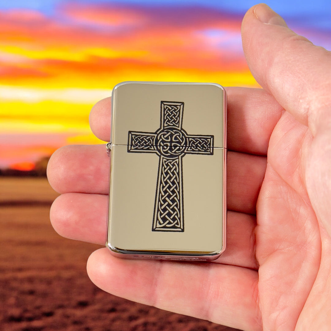 Hand holding a HipFlaskPlus Celtic cross chrome lighter at sunset