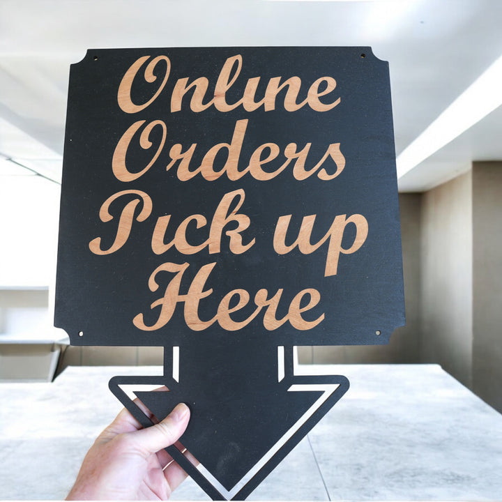 Hand holding a HipFlaskPlus Online Orders Pick Up Here black wood arrow sign in a parking lot