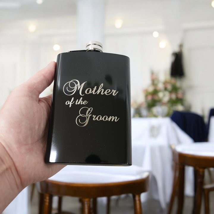 Hand holding a HipFlaskPlus 8oz black stainless steel hip flask engraved with Mother of the Groom in a room