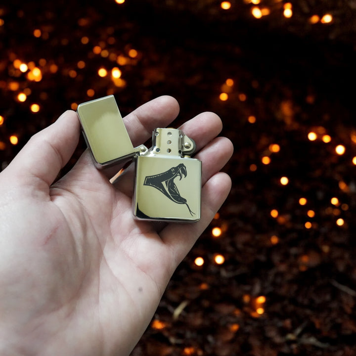 Hand holding an open HipFlaskPlus poison viper lighter with warm bokeh lights