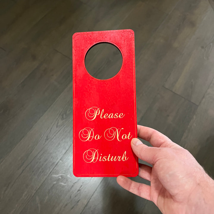 Hand holding a HipFlaskPlus red Please Do Not Disturb wood door hanger on a gray background
