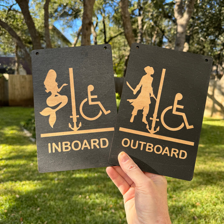 Hand holding a HipFlaskPlus Restrooms Inboard and Outboard black wood signs outdoors