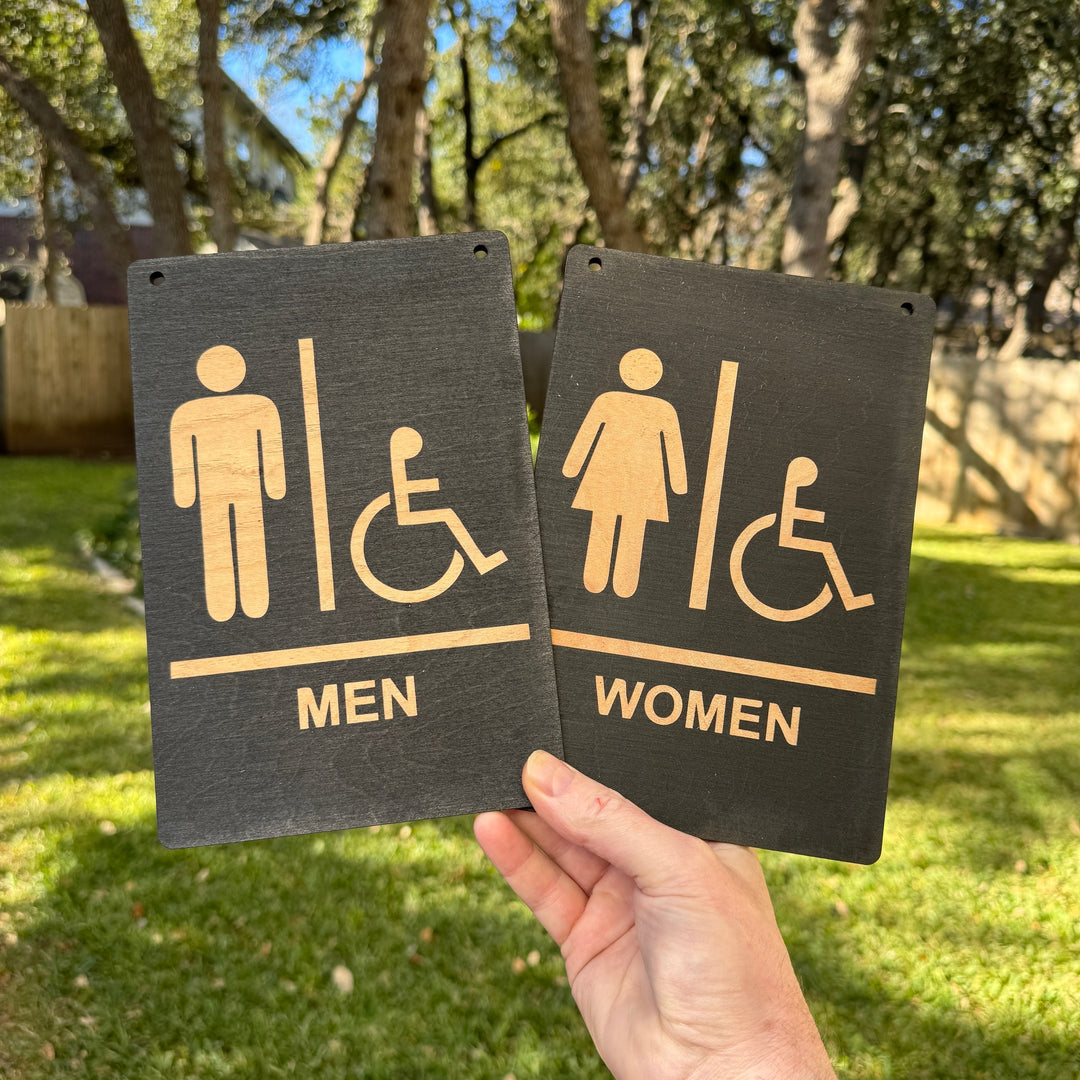 Hand holding a HipFlaskPlus Restrooms Men and Women black wood signs outdoors