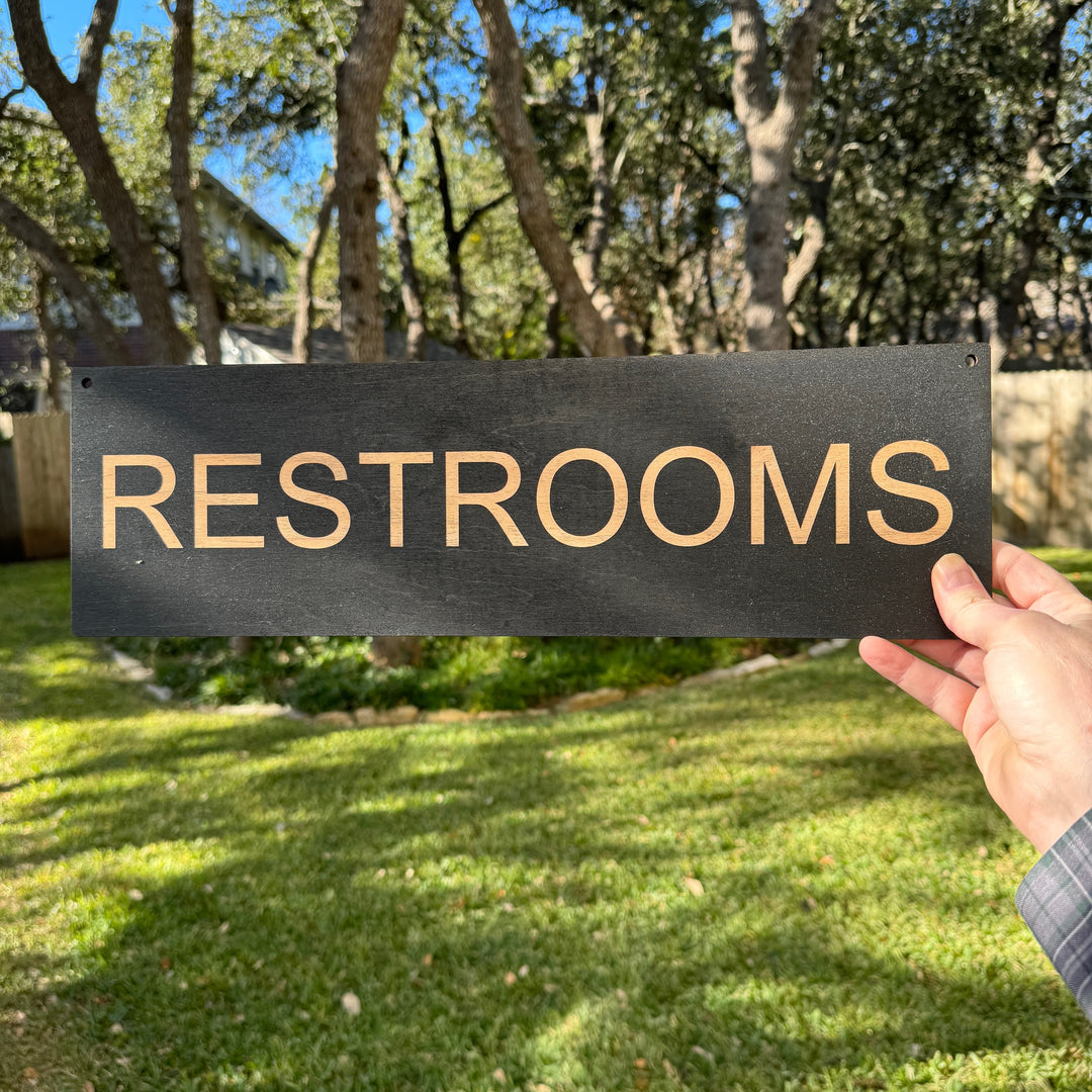 Hand holding a HipFlaskPlus black wood Restrooms sign outdoors